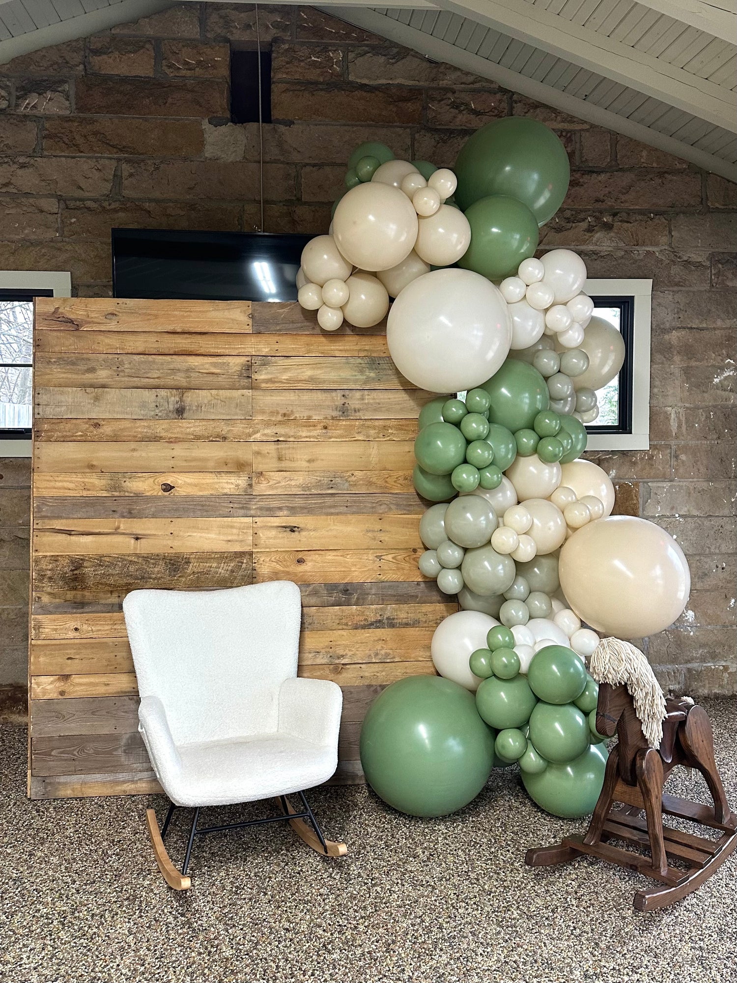 Balloon garland on wooden backdrop with green, cream, and white balloons with a white chair and wooden horse.